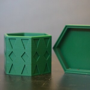 Könnte beinhalten: Eine grüne sechseckige Box mit einem geometrischen Muster an den Seiten. Der Deckel ist geöffnet und die Box steht auf einer dunklen Oberfläche.