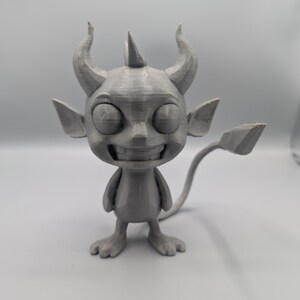 Puede incluir: Figura gris impresa en 3D de un diablo de dibujos animados con cuernos, orejas puntiagudas y una cola. La figura tiene una amplia sonrisa y ojos grandes y redondos. El diablo está de pie.