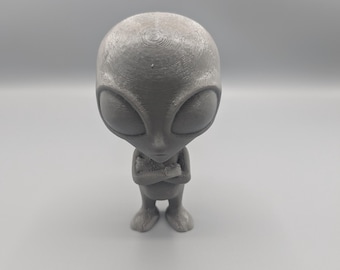 Puede incluir: Una figura de alienígena gris con una cabeza grande, ojos grandes y los brazos cruzados sobre el pecho. La figura está de pie y tiene pies pequeños. El alienígena es de color gris claro y parece estar hecho de un material sólido.
