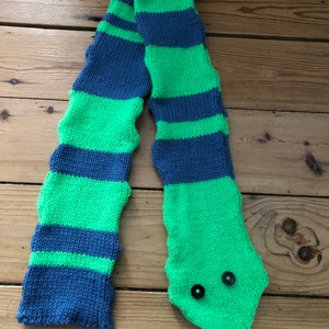 Puede incluir: Una bufanda de punto a rayas verdes y azules con un diseño de cabeza de serpiente. La cabeza de serpiente tiene dos botones negros para los ojos.