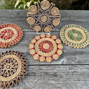 May include: Five circular woven coasters featuring intricate patterns in tan combined with contrasting colours including dark blue, red, green, and brown. Each coaster has a textured, coiled design with a scalloped edge and a central floral motif.