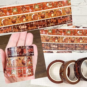 May include: Washi tape rolls with a food theme. The tape features illustrations of baked goods, mushrooms, and the words "Pie Bread Cake Cookies". The colour palette includes shades of brown, orange, and white. The image shows the tape rolls and a strip of the tape.
