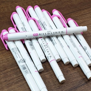 Puede incluir: Una pila de rotuladores de doble punta Zebra Mildliner rosa y blanco. Los rotuladores tienen una tapa rosa y un cuerpo blanco con el texto "ZEBRA Midink MILDLINER Double Ended" impreso en el cuerpo.