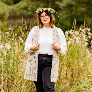 May include: A woman wearing a beige crochet vest with a white shirt and black jeans. She is wearing a flower crown and glasses. The background is a field of tall grass.