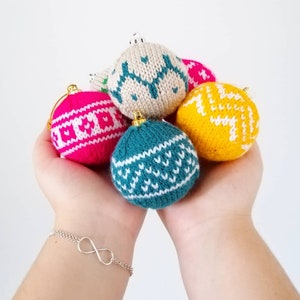 Könnte beinhalten: Eine Sammlung von sechs gestrickten Weihnachtskugeln in verschiedenen Farben und Mustern, darunter rosa, weiß, türkis, gelb und beige. Die Kugeln werden in den Händen einer Person gehalten.