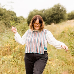 May include: A woman wearing a colorful crocheted vest with a white blouse and black jeans. She is standing in a field of tall grass and wildflowers.