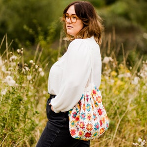 Könnte beinhalten: Eine Frau in einer weißen Bluse und schwarzen Jeans trägt eine gehäkelte Tasche mit einem bunten Granny-Square-Muster. Die Tasche hat einen langen Riemen und hängt an ihrer Schulter.