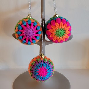 K&ouml;nnte beinhalten: Drei geh&auml;kelte Weihnachtskugeln in verschiedenen Farben, darunter Pink, Orange, Gr&uuml;n und Blau. Jede Kugel hat ein Sternmuster in der Mitte.