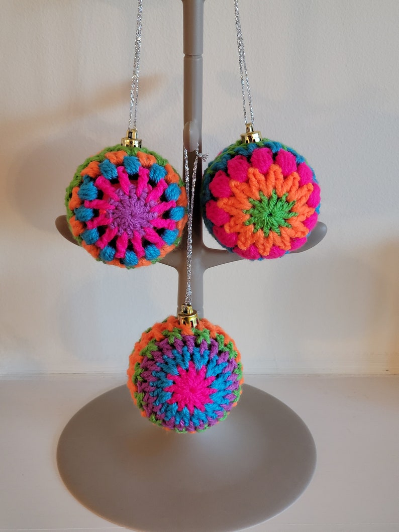 K&ouml;nnte beinhalten: Drei geh&auml;kelte Weihnachtskugeln in verschiedenen Farben, darunter Pink, Orange, Gr&uuml;n und Blau. Jede Kugel hat ein Sternmuster in der Mitte.