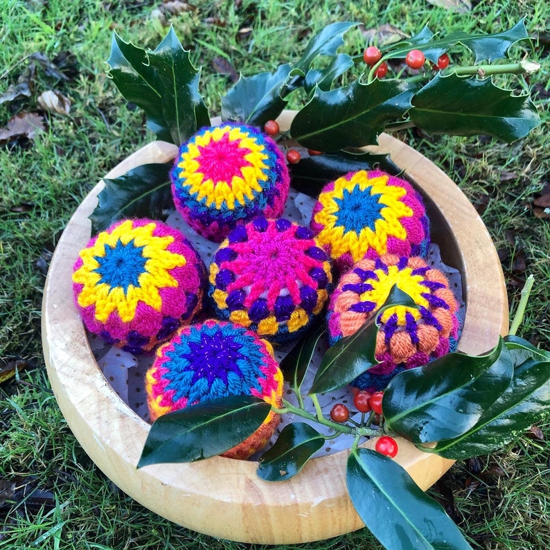 K&ouml;nnte beinhalten: Sechs bunte geh&auml;kelte Ornamente in einer Holzschale. Die Ornamente sind rosa, gelb, blau, lila und orange. Die Schale ist von gr&uuml;nen Stechpalmenbl&auml;ttern umgeben.