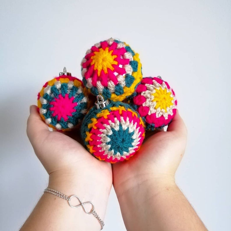 K&ouml;nnte beinhalten: Vier geh&auml;kelte Weihnachtskugeln in Rosa, Gelb und T&uuml;rkis auf wei&szlig;em Hintergrund. Die Kugeln sind kugelf&ouml;rmig und haben ein geometrisches Muster.
