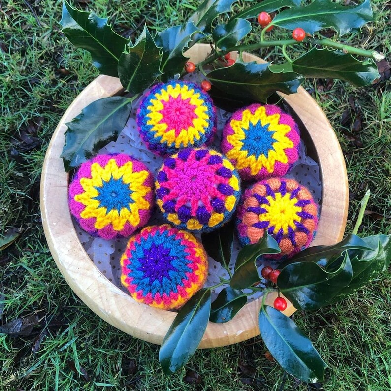 K&ouml;nnte beinhalten: Sechs bunte geh&auml;kelte Ornamente in einer Holzschale. Die Ornamente sind rosa, gelb, blau und lila. Die Schale ist von gr&uuml;nen Stechpalmenbl&auml;ttern umgeben.