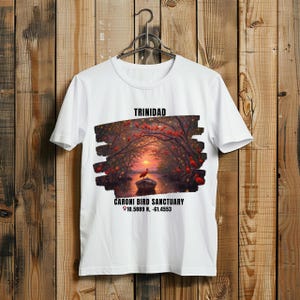 May include: White t-shirt featuring a scenic illustration of the Caroni Bird Sanctuary in Trinidad. The design includes a sunset, birds, and the text "TRINIDAD" and "CARONI BIRD SANCTUARY". Coordinates are also printed on the shirt.