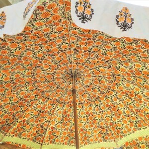 K&ouml;nnte beinhalten: Ein gro&szlig;er Sonnenschirm mit einem dichten, sich wiederholenden Muster aus orangefarbenen und gelben Blumenmotiven auf hellem Grund. Der &auml;u&szlig;ere Rand zeigt eine wei&szlig;e Borte mit einzelnen, orangefarbenen und schwarzen Blumenmustern.