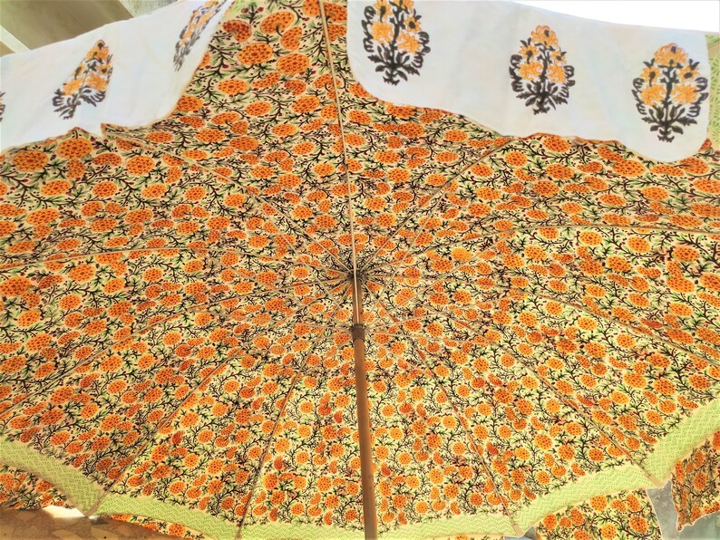 K&ouml;nnte beinhalten: Ein gro&szlig;er Sonnenschirm mit einem dichten, sich wiederholenden Muster aus orangefarbenen und gelben Blumenmotiven auf hellem Grund. Der &auml;u&szlig;ere Rand zeigt eine wei&szlig;e Borte mit einzelnen, orangefarbenen und schwarzen Blumenmustern.