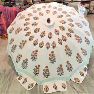 K&ouml;nnte beinhalten: Ein wei&szlig;er Sonnenschirm mit einem sich wiederholenden Muster aus orangefarbenen und schwarzen Blumenmotiven in konzentrischen Kreisen. Der Schirm hat einen hellgr&uuml;nen Zickzack-Rand und einen dunkelbraunen Holzknauf an der Spitze.