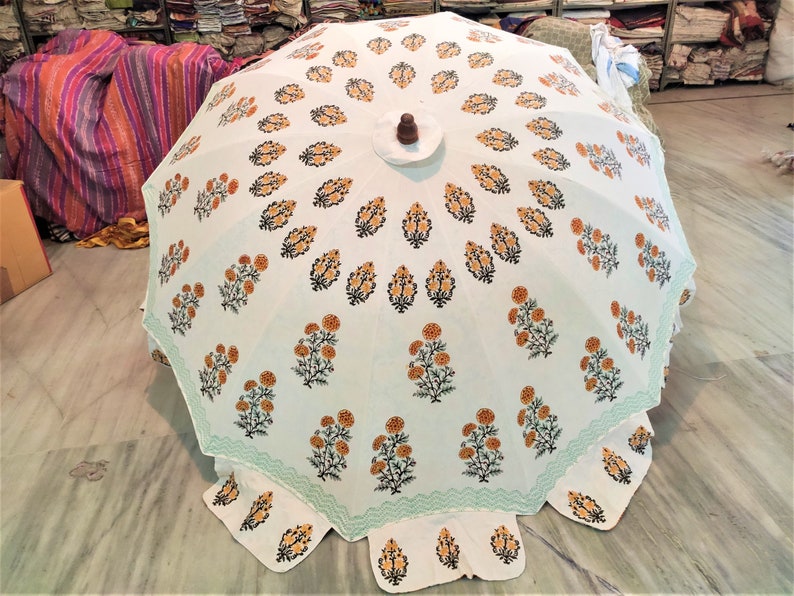 K&ouml;nnte beinhalten: Ein wei&szlig;er Sonnenschirm mit einem sich wiederholenden Muster aus orangefarbenen und schwarzen Blumenmotiven in konzentrischen Kreisen. Der Schirm hat einen hellgr&uuml;nen Zickzack-Rand und einen dunkelbraunen Holzknauf an der Spitze.