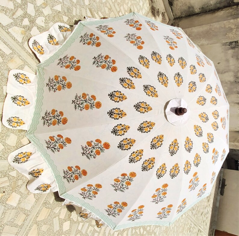 K&ouml;nnte beinhalten: Ein wei&szlig;er Sonnenschirm mit einem sich wiederholenden Blumenmuster aus orangefarbenen Ringelblumen mit gr&uuml;nen Stielen und Bl&auml;ttern. Der Rand ist mit einer hellgr&uuml;nen Bogenkante versehen, und in der Mitte befindet sich ein dunkelbrauner Holzknauf.