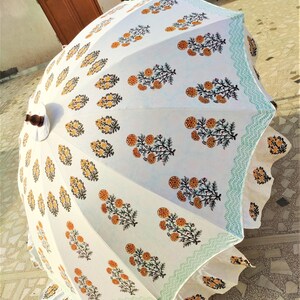 K&ouml;nnte beinhalten: Ein wei&szlig;er Sonnenschirm mit einem sich wiederholenden Blumenmuster aus orangefarbenen Ringelblumen mit gr&uuml;nen Stielen und Bl&auml;ttern. Die R&auml;nder sind mit einer hellblauen geometrischen Borte und einem ger&uuml;schten Stoffsaum verziert.