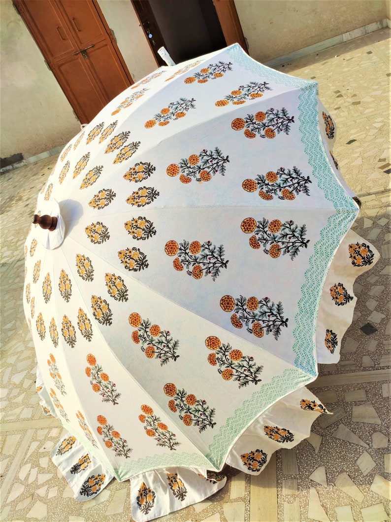 K&ouml;nnte beinhalten: Ein wei&szlig;er Sonnenschirm mit einem sich wiederholenden Blumenmuster aus orangefarbenen Ringelblumen mit gr&uuml;nen Stielen und Bl&auml;ttern. Die R&auml;nder sind mit einer hellblauen geometrischen Borte und einem ger&uuml;schten Stoffsaum verziert.