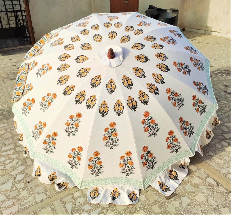 K&ouml;nnte beinhalten: Ein gro&szlig;er wei&szlig;er Sonnenschirm mit einem sich wiederholenden Blumenmuster aus orangefarbenen und gelben, ringelblumenartigen Bl&uuml;ten mit gr&uuml;nen Stielen. Der Schirm hat einen gewellten Rand mit hellgr&uuml;ner Zickzack-Borte und einen dunkelbraunen Holzknauf.