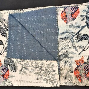May include: A white cotton quilt with a blue and white floral pattern. The quilt features a repeating pattern of owls and branches. The quilt is folded in half and has a blue backing.