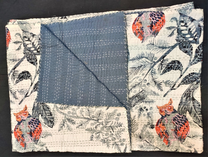 May include: A white cotton quilt with a blue and white floral pattern. The quilt features a repeating pattern of owls and branches. The quilt is folded in half and has a blue backing.