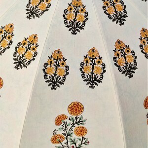 K&ouml;nnte beinhalten: Eine hellfarbige Stoffoberfl&auml;che mit einem sich wiederholenden Muster aus gedruckten Blumenmotiven. Das Design zeigt gelbe und orangefarbene Bl&uuml;ten mit schwarzen Stielen und Bl&auml;ttern, die in vertikalen Abschnitten durch feine N&auml;hte getrennt sind.