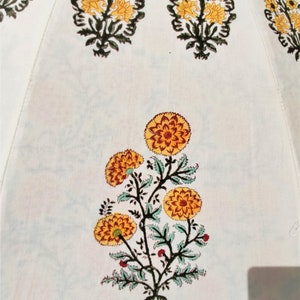 K&ouml;nnte beinhalten: Eine wei&szlig;e Stoffoberfl&auml;che mit einem sich wiederholenden, blockgedruckten Blumenmuster. Das Design besteht aus stilisierten gelben und orangefarbenen, ringelblumenartigen Bl&uuml;ten mit dunkelgr&uuml;nen Stielen und Bl&auml;ttern, die symmetrisch angeordnet sind.