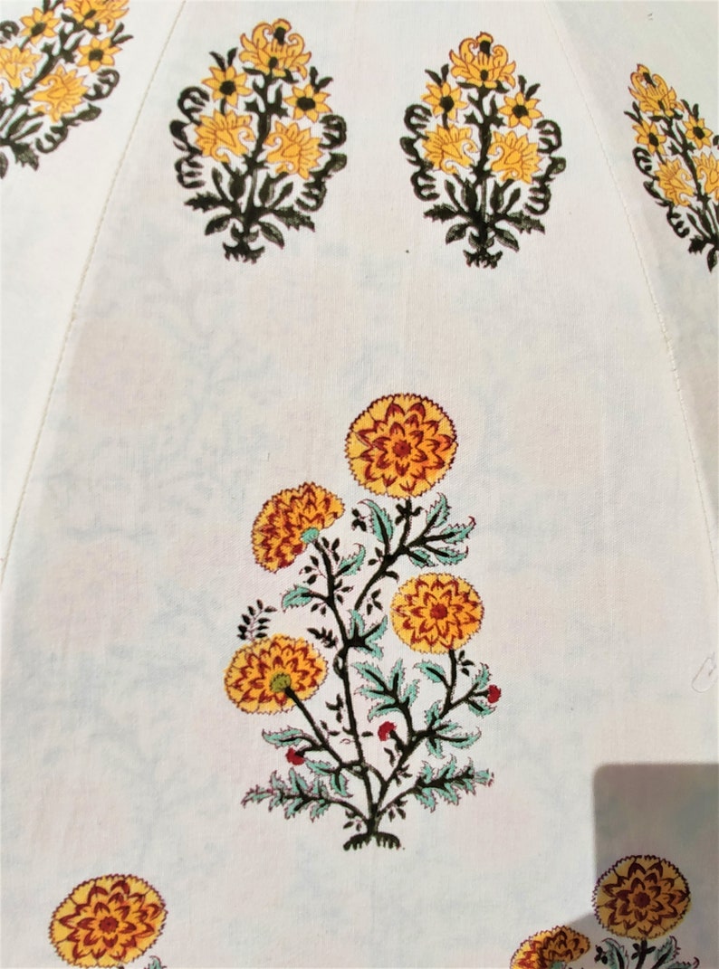 K&ouml;nnte beinhalten: Eine wei&szlig;e Stoffoberfl&auml;che mit einem sich wiederholenden, blockgedruckten Blumenmuster. Das Design besteht aus stilisierten gelben und orangefarbenen, ringelblumenartigen Bl&uuml;ten mit dunkelgr&uuml;nen Stielen und Bl&auml;ttern, die symmetrisch angeordnet sind.