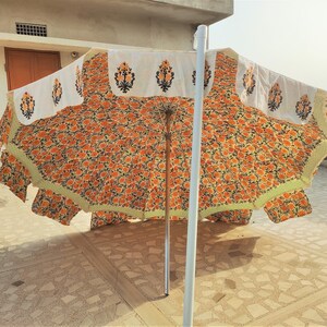 K&ouml;nnte beinhalten: Ein gro&szlig;er Sonnenschirm f&uuml;r den Au&szlig;enbereich mit einem lebendigen orange-schwarzen Blumenmuster auf dem Schirm. Der &auml;u&szlig;ere Rand ist mit einer wei&szlig;en Borte verziert, die einzelne schwarze und orange Blumenmotive zeigt und hellgr&uuml;n ges&auml;umt ist.
