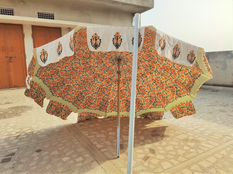 K&ouml;nnte beinhalten: Ein gro&szlig;er Sonnenschirm f&uuml;r den Au&szlig;enbereich mit einem lebendigen orange-schwarzen Blumenmuster auf dem Schirm. Der &auml;u&szlig;ere Rand ist mit einer wei&szlig;en Borte verziert, die einzelne schwarze und orange Blumenmotive zeigt und hellgr&uuml;n ges&auml;umt ist.