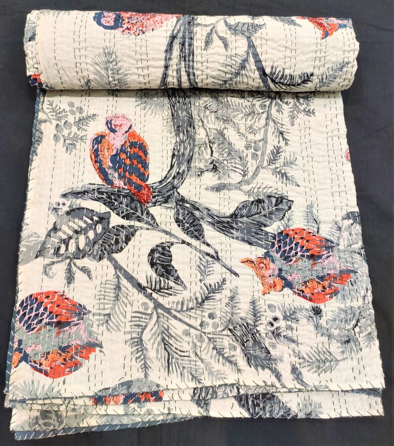 May include: A white kantha quilt with a black and gray floral pattern and red and pink birds. The quilt is folded and rolled up.