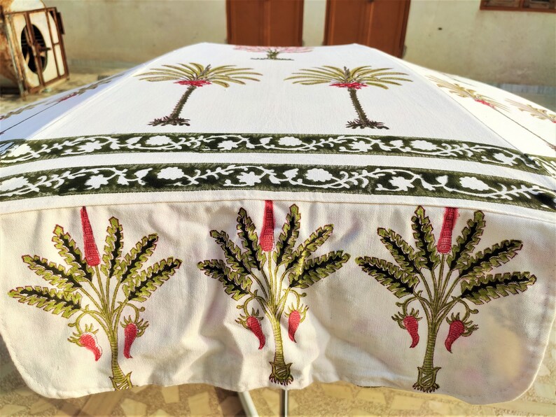 May include: White cotton umbrella with a green and red floral print. The umbrella has a double border of green and white floral designs. The umbrella is closed and sitting on a table.