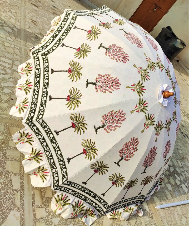 May include: A white cotton umbrella with a green and pink floral print. The umbrella has a scalloped edge and a wooden handle.