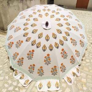 K&ouml;nnte beinhalten: Ein wei&szlig;er Sonnenschirm mit einem sich wiederholenden Muster aus gelben und gr&uuml;nen Blumenmotiven. Der Schirm hat einen gewellten Rand mit einer hellgr&uuml;nen Bord&uuml;re und einen dunkelbraunen Holzknauf in der Mitte. Der Stoff zeigt ein symmetrisches Design.