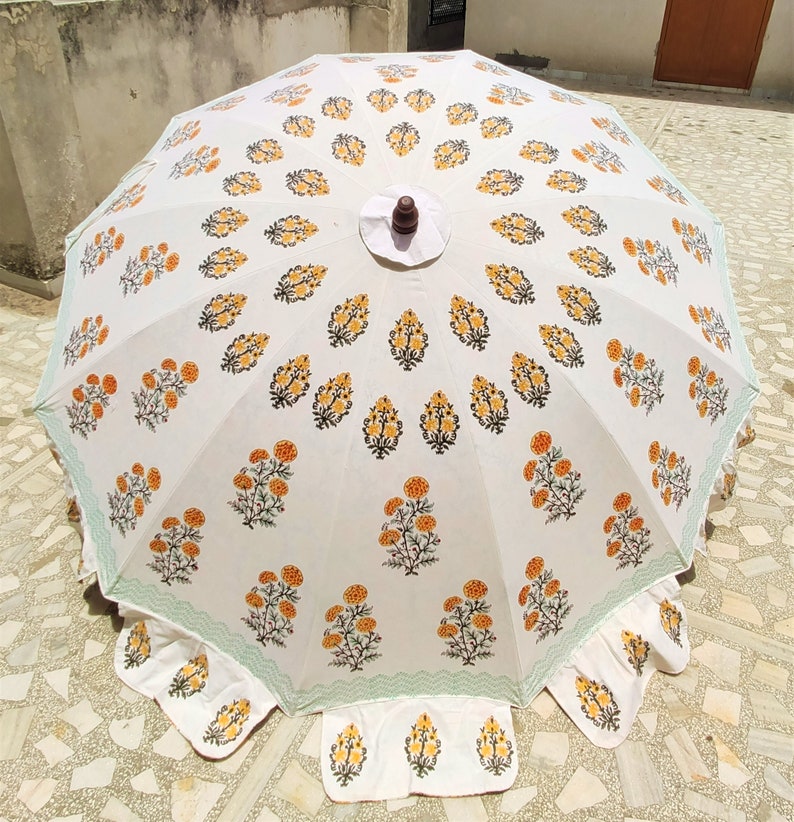 K&ouml;nnte beinhalten: Ein wei&szlig;er Sonnenschirm mit einem sich wiederholenden Muster aus gelben und gr&uuml;nen Blumenmotiven. Der Schirm hat einen gewellten Rand mit einer hellgr&uuml;nen Bord&uuml;re und einen dunkelbraunen Holzknauf in der Mitte. Der Stoff zeigt ein symmetrisches Design.