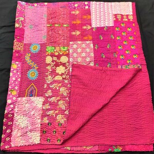 May include: A rectangular patchwork quilt featuring a vibrant magenta base color. The surface is composed of various fabric squares with diverse floral, geometric, and paisley patterns in shades of pink, yellow, green, blue, and white, all joined by visible running stitch quilting.