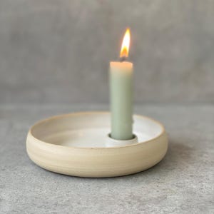 Op de afbeelding: Een brandende groene kaars in een beige keramische kandelaar. De kandelaar heeft een cirkelvormig ontwerp met een witte binnenkant en een beige buitenkant. De vlam is heldergeel en oranje.