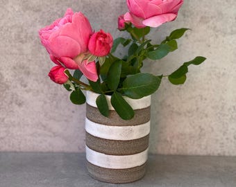 Può includere: Un vaso di ceramica a strisce orizzontali marroni e bianche contiene un bouquet di rose rosa e foglie verdi. Le rose sono in piena fioritura, con diversi boccioli. Il vaso è cilindrico e poggia su una superficie grigia.