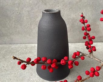 Könnte beinhalten: Eine mattschwarze Keramikvase mit leicht strukturierter Oberfläche. Die Vase ist zylindrisch mit rundem Boden und schmaler Öffnung. Rote Beeren an einem Zweig sind in der Nähe der Vase angeordnet.