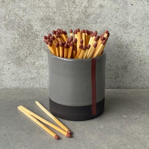 Könnte beinhalten: Ein grauer Keramik-Streichholzhalter mit einem schwarzen und burgunderfarbenen Streifenmuster. Der Halter ist mit Streichhölzern aus Holz mit roten Köpfen gefüllt.