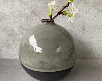 Könnte beinhalten: Eine zweifarbige Keramikvase mit kugelförmiger Form. Die obere Hälfte ist ein gedämpftes Grün, während die untere Hälfte schwarz ist. Ein kleiner Zweig mit weißen Blüten und grünen Blättern befindet sich in der Vase und bildet ein minimalistisches Blumenarrangement.