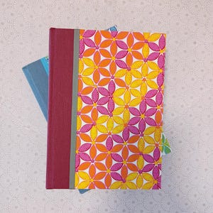 May include: A pink and orange floral patterned notebook with a burgundy spine and a grey ribbon bookmark. The notebook is stacked on top of a blue notebook.