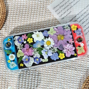 May include: A clear Switch console case filled with colorful dried flowers. The case has blue and red accents on the sides. The flowers include daisies, pansies, and other blooms in shades of purple, yellow, white, and pink. A handwritten note is in the background.