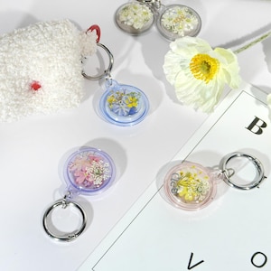 May include: A collection of keychains with dried flowers encased in clear resin. The keychains have silver-colored rings and are in various colors, including blue, pink, and clear. A white fluffy pouch with a red accent is also present.
