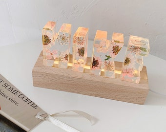 May include: A wooden base with a light switch holds five clear resin letters spelling "MIKE" with dried flowers pressed inside. The letters are illuminated from behind, casting a warm glow.