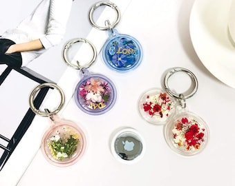 May include: A collection of keychains with clear, circular pendants containing dried flowers and decorative elements. Each keychain has a silver-colored ring. One keychain has the word "Love" in blue. The image also includes an Apple AirTag.