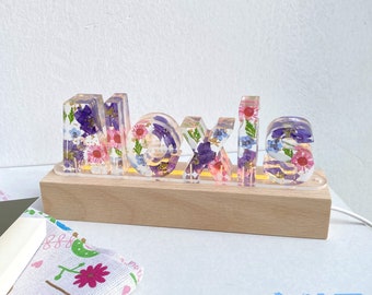 May include: A custom name sign with the name "Moxie" spelled out in clear resin letters. The letters are filled with pressed flowers in shades of pink, purple, and blue. The sign is illuminated from behind with a warm white light and sits on a wooden base. The sign is made by Cute Secret Garden.