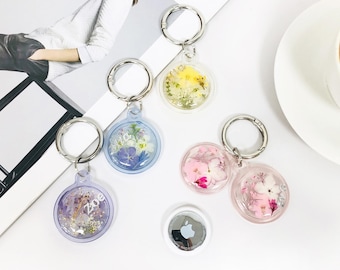 May include: Five keychains with clear resin charms containing pressed flowers. The charms are attached to silver key rings. The charms are in various colors, including purple, pink, blue, and yellow. The keychains are arranged on a white surface with a magazine and a cup of coffee in the background.